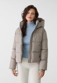 Bundička so zapínaním na zips v svetlej béžovej farbe, s kapucňou a prešívaným dizajnom. Pod ňou je vidieť svetlomodrý rebrovaný rolák.