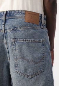 Denim jeans with a light blue wash, featuring a brown leather patch at the back waist and a subtle curved stitching detail on the pocket.