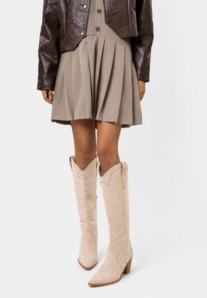 Kvinde iført beige plisseret nederdel, cropped mørkebrun læderjakke og knæhøje lysebeige ruskinds cowboystøvler med hæle.