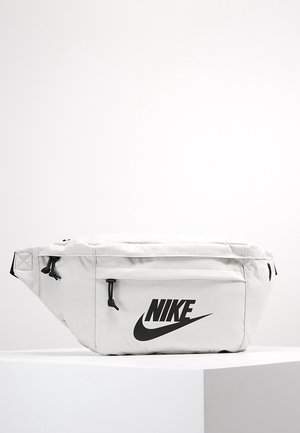 Witte Nike heuptas met zwart logo en ritstrekker, rustend op een wit voetstuk tegen een effen lichte achtergrond.