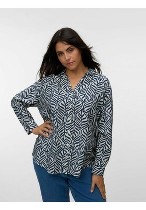 Tumejate juustega naine kannab valget pika varrukaga särki tumedasiniste lehemustriga ja siniseid teksapükse, poseerides käega puusal ühevärvilise tausta ees.