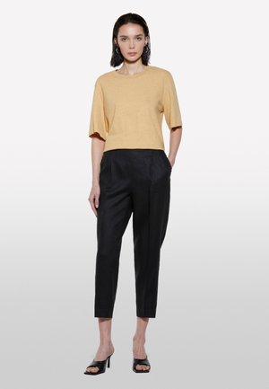 Top giallo a maniche corte con vestibilità rilassata abbinato a pantaloni scuri e appuntiti. Il modello indossa sandali neri con tacco aperti. Texture di tessuto lisce.