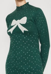 Grön stickad tröja med hög krage, prydd med en vit rosettgrafik och silverprickar genom hela tyget. Långa ärmar, figurnära passform.