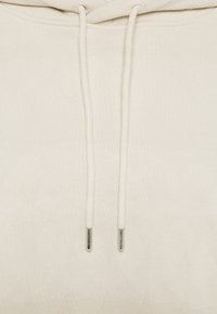 Sudadera beige con una tela texturizada, que cuenta con una capucha con cordón y puntas metálicas plateadas, además de un diseño simple.