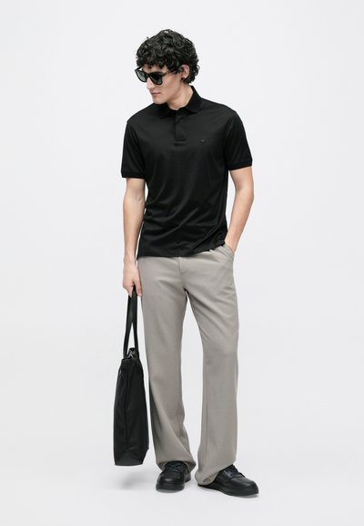 Melns polo krekls ar īsām piedurknēm, pelēkas bikses un melni sporta apavi. Modelis tur melnu rokas somu un valkā tumšas saulesbrilles.