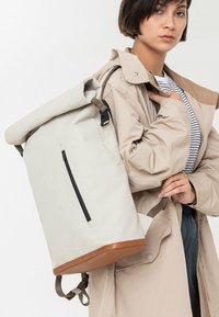 Roll-top rugzak in lichtgrijze stof met een bruine basis. Voorzien van een voorvak met ritssluiting en verstelbare bandjes voor veilig dragen.