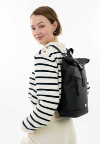 Sac à dos noir à rabat avec poches zippées et bretelles réglables, fabriqué à partir de matériaux durables. Le modèle porte un pull rayé.