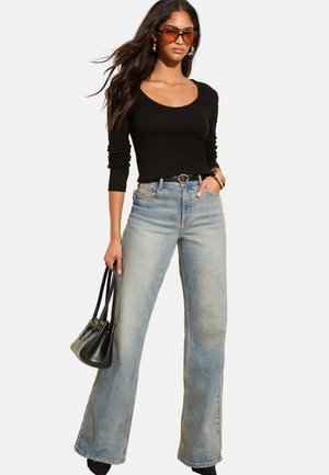 Mujer con gafas de sol ámbar, camiseta negra de manga larga, vaqueros acampanados de tiro alto azul claro, cinturón negro, sosteniendo un bolso negro.