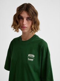 Młoda osoba z krótkimi falistymi brązowymi włosami, ubrana w ciemnozieloną sportową koszulkę hummel z białym logo na piersi.