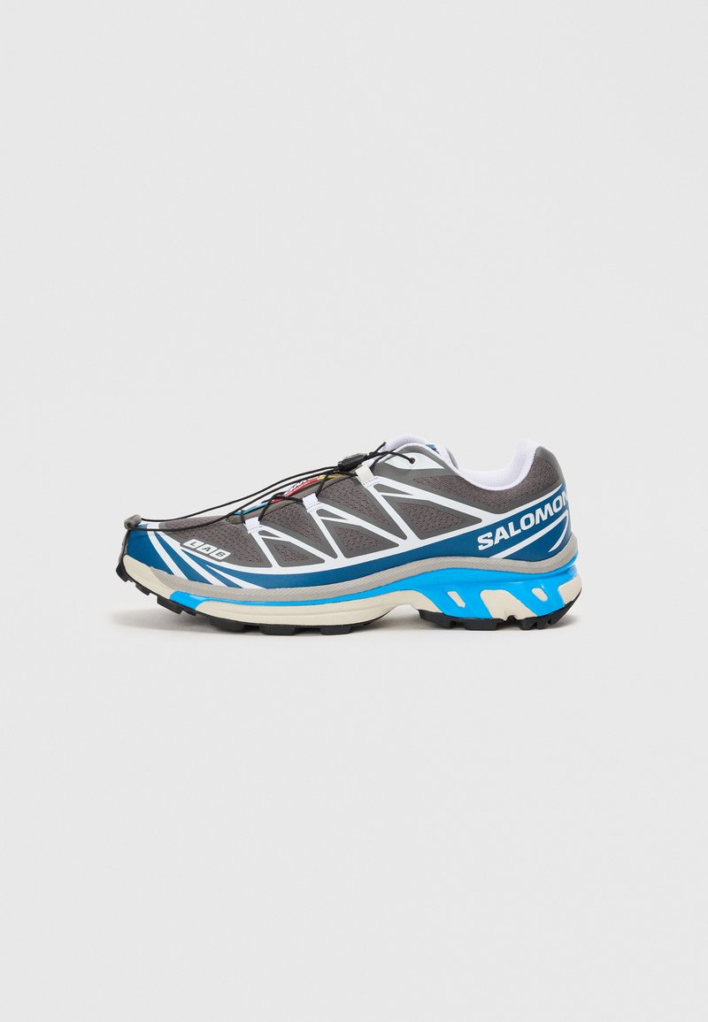 Sive in modri Salomon čevlji za trail tek z belimi detajli, črnimi hitrimi vezalkami in robustnim podplatom na svetli podlagi.