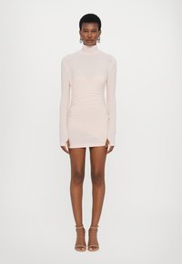 Langmouw, bodycon mini-jurk van zacht roze stof. Heeft een hoge halslijn en geplooide details aan de zijkant. Gecombineerd met bandjeshakken.
