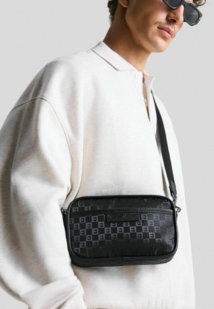 Sac en bandoulière noir à motifs avec fermeture éclair, présentant une surface texturée, une sangle ajustable et une forme rectangulaire, porté sur un sweatshirt blanc.