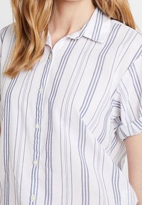 Camicia leggera button-up bianca con righe verticali blu, maniche corte arrotolate e colletto classico. Presenta bottoni perlacei.