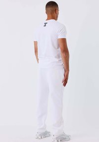 Uomo di spalle che indossa una maglietta bianca con la scritta "CAMBIA IL GIOCO" sulla parte superiore della schiena, pantaloni della tuta bianchi e sneakers bianchi.