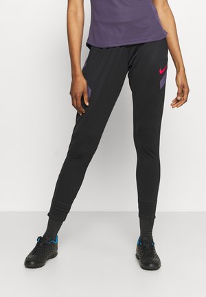 Person trägt schwarze Sporthose mit lila Akzenten und rotem Nike-Logo, kombiniert mit schwarzen Schuhen und einem lila Oberteil.