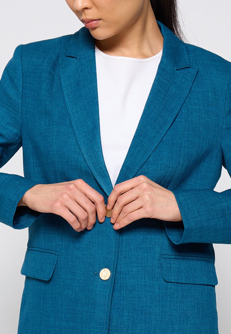 Blazer turquoise en tissu texturé, avec une fermeture à un seul bouton en bois et deux poches avant. Le modèle est en train de fermer le blazer.
