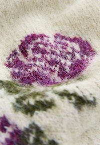 Tissu en tricot crème avec une texture duveteuse présentant un motif de fleurs tricotées en violet et blanc avec des feuilles vertes.
