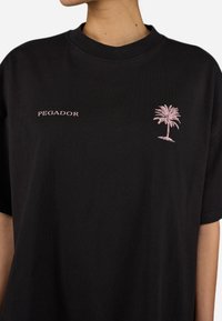 Camiseta de algodón negra con un diseño de palmera rosa bordado y el nombre de la marca "PEGADOR" arriba. Cuello redondo y mangas cortas.