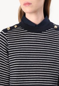 Pulover striped în navy și alb, cu un guler ribalit și o cămașă ascunsă cu buton la gât dedesubt, având butoane aurii pe umeri.