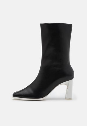 Bottine en cuir noir avec un bout pointu et un talon bloc blanc. Texture lisse et design minimaliste, adaptée à diverses occasions.