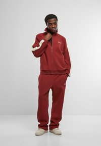 Bordový teplákový komplet s krémovými akcentmi; sako na zips a zodpovedajúce nohavice; mäkká tkanina, priliehavý dizajn, logo na hrudi a na bokoch.