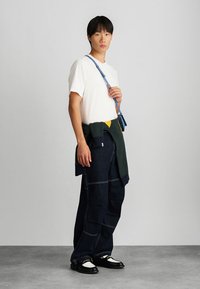 Camiseta blanca, pantalones vaqueros de pierna ancha de color azul oscuro con costuras visibles, combinados con un suéter verde anudado y un bolso de tirantes azul. Zapatos en blanco y negro.