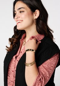 Zwarte gewatteerde vest over een roze kant blouse, met ronde gouden creolen en drie gouden armbanden om de pols.