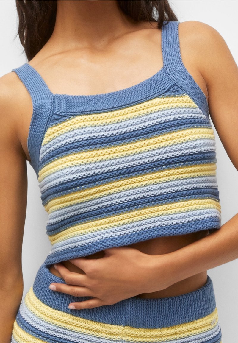 Žena v bezrukávovém pleteném crop topu s horizontálními pruhy v modré, žluté a bílé barvě, v kombinaci se shodnými kalhotami.