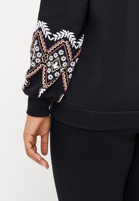 Svart sweatshirt med blommig och geometrisk broderi i vitt, rosa och gult på ärmarna. Ribbad mudd och nederkant. Mjuk textur.