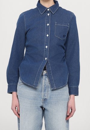 Person trägt ein eng anliegendes, dunkelblaues Denim-Hemd mit Brusttasche und hellblaue, hoch taillierte Jeans, steht vor einfachem Hintergrund.