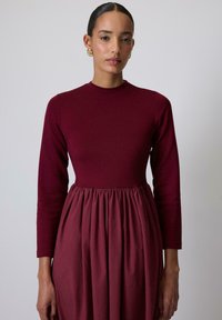 Vestido burdeos con una parte superior de manga larga ajustada y una falda agrupada y fluida. Tejido suave con una textura sutil, escote redondo.