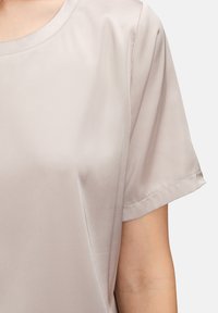 Satijnen top met korte mouwen in zacht beige, voorzien van een ronde halslijn en een gladde textuur met een subtiele glans; naden zichtbaar bij de schouders en de zoom.