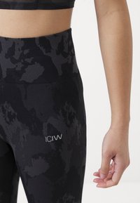 Zwarte leggings met een camouflagedessin, hoge tailleband, gestructureerde stof en ICIW-logo op de linkerheup; close-up van een hand die ernaast rust.