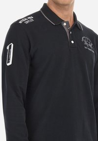 Polo shirt nero a maniche lunghe con colletto a contrasto. Presenta un logo ricamato e il numero "1" sulla manica. Realizzato in tessuto di cotone.
