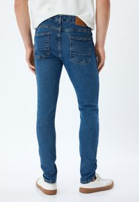 Jeans de denim azul ajustados con un acabado texturizado, diseño de cinco bolsillos y parche de cuero marrón en la cinturilla. Combinados con zapatillas blancas.
