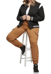 Svart och vit collegejacka, tan cargo-byxor med elastiska muddar, och svarta höga sneakers. Materialet verkar mjukt med en avslappnad design.