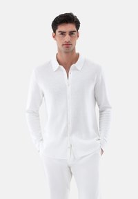 Weißes Langarmhemd aus strukturiertem Mesh-Gewebe, mit einem Button-Down-Kragen und Frontknöpfen, kombiniert mit weißen Hosen.