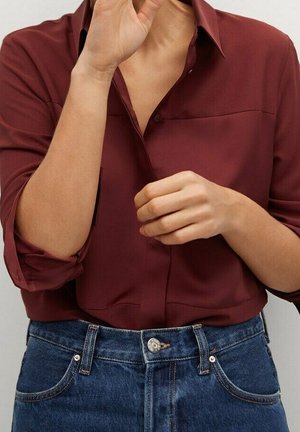 Personne portant une chemise bordeaux boutonnée et un jean bleu avec les manches retroussées, ajustant son col d'une main.