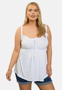 Valkoinen hihaton toppi, jossa on röyhelökaulus ja säädettävä nauha. Pehmeä kangas laskeutuu vartalon ylle, ja se on yhdistetty tummiin farkkushortseihin.