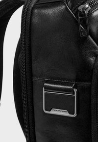 Sac en cuir noir avec une finition texturée, un accent en métal argenté et une fermeture éclair. Présente une plaque métallique de marque sur le côté.