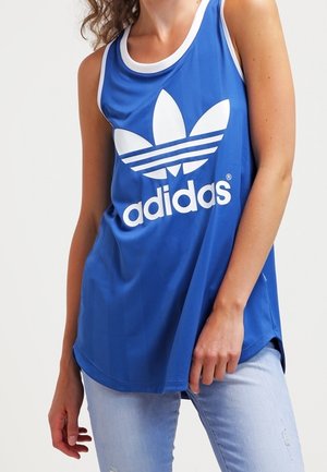Woman wearing a blue Adidas sleeveless top with white logo and light blue ripped jeans, posing with one hand near waist.
