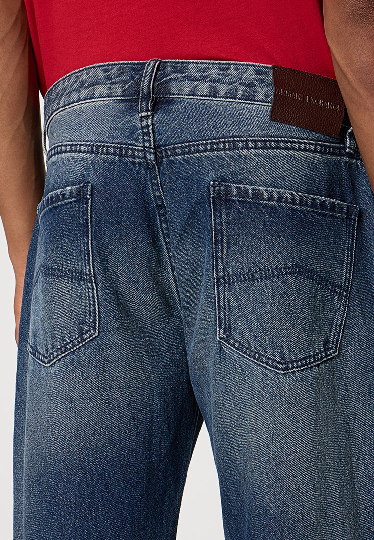 Dżinsy denimowe z dwoma tylnymi kieszeniami, wzorzystym pasem i brązową skórzaną metką Armani Exchange, noszone z czerwoną koszulą.