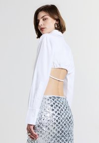 Camicia bianca con bottoni, con retro corto e dettagli elasticizzati. Abbinata a una gonna di paillettes argentate con un motivo testurizzato.