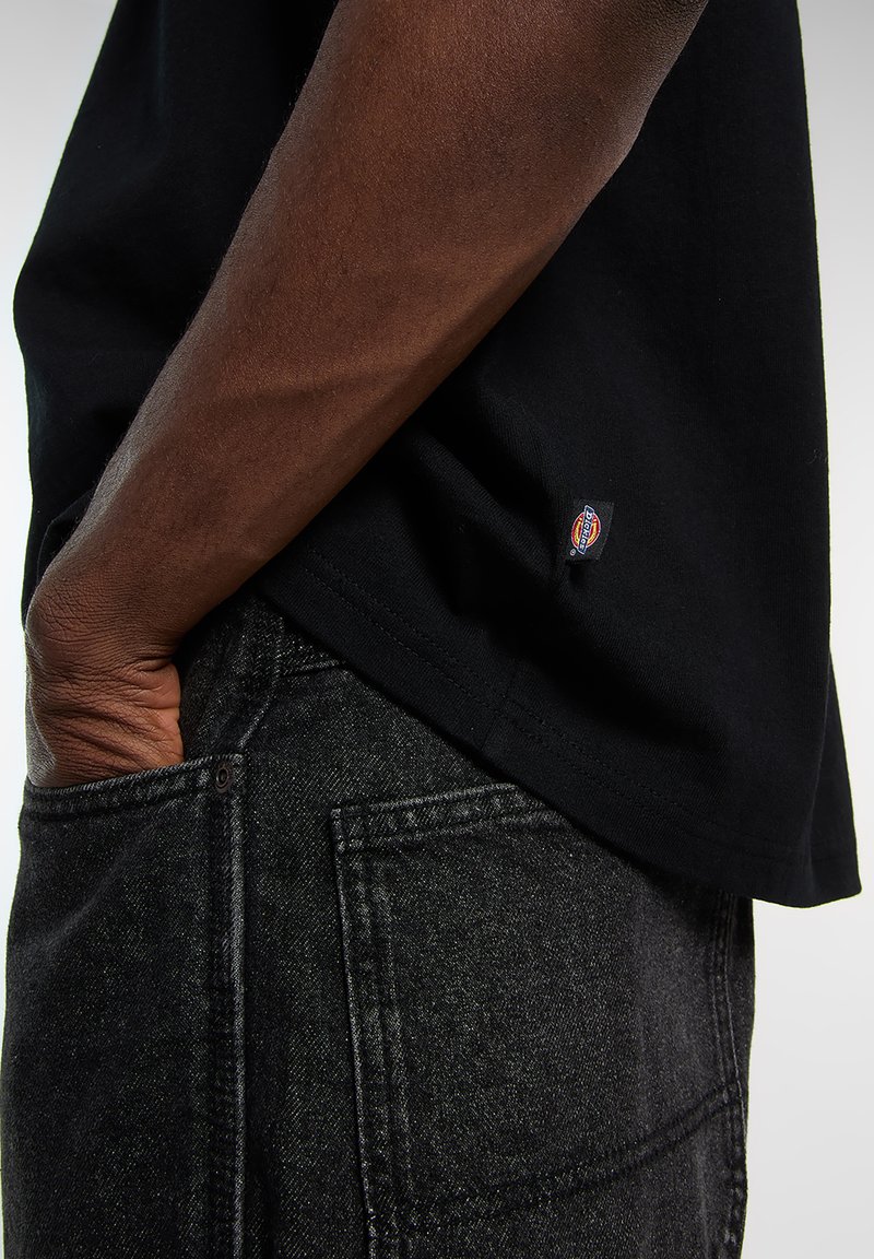 Black cotton t-shirt with a straight hem, featuring a small logo tag at the bottom. Worn with dark denim jeans, showing textured fabric.