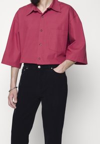 Červená krátká košile s knoflíky, špičatým límečkem a jednou náprsní kapsou, doplněná černými manšestrovými kalhotami s předními kapsami.