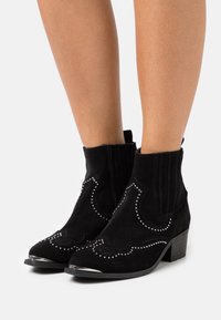 Botas de tobillo de ante negro con adornos de tachuelas plateadas, punta afilada, tacón bajo y paneles laterales elásticos. Diseño texturizado y puntera brillante.