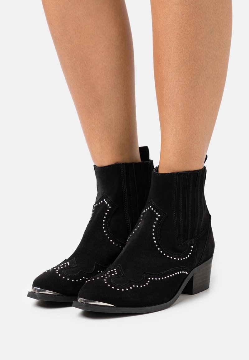 Botas de tobillo de ante negro con adornos de tachuelas plateadas, punta afilada, tacón bajo y paneles laterales elásticos. Diseño texturizado y puntera brillante.