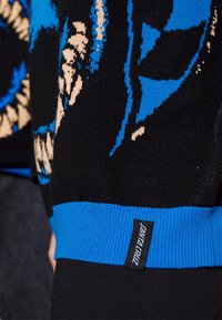 Svart tröja med blå och beige grafiska mönster, ribbad blå mudd och en 'Santa Cruz' etikett på sidan.