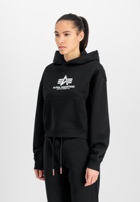 Čierna skrátená mikina s kapucňou vyrobená z bavlnenej zmesi, s predným vreckom, s bielym logom "ALPHA INDUSTRIES" a dizajnom horizontálnych čiar.