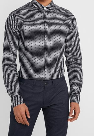 Mand iført sort og hvidt stribet skjorte med knapper og mørkeblå bukser, stående med hænderne afslappet langs siderne.